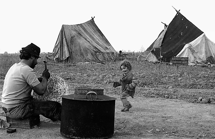 Roemenie 1992. Tijdelijk kamp van de Calderari zigeuners.  Een man repareert een ketel en zijn dochterje danst op het ritme van de hamerslagen.
